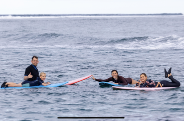 Quatre personnes, dont un enfant, sont allongées sur des planches de surf dans l'océan, souriantes et se tenant la main, profitant ensemble d'une journée agréable et détendue sur l'eau.
