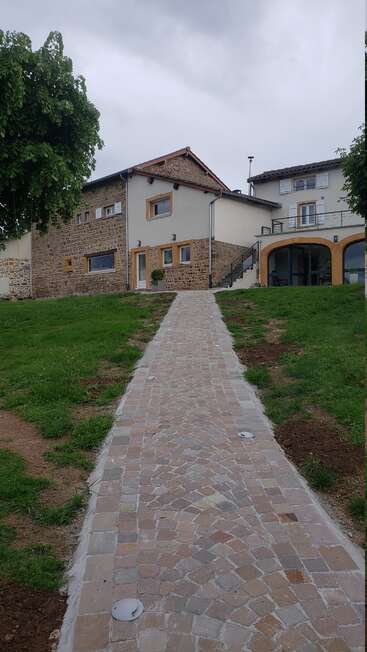 Ein Steinweg führt zu einem hübschen, modern renovierten Haus mit rustikalen Ziegeln und weißen Wänden, umgeben von Gras, unter einem bewölkten Himmel und eingerahmt von Bäumen.