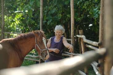 Une femme âgée sourit dans un espace clôturé en bois, avec un cheval brun à proximité. Un feuillage vert luxuriant les entoure, créant une scène paisible et naturelle en plein air.