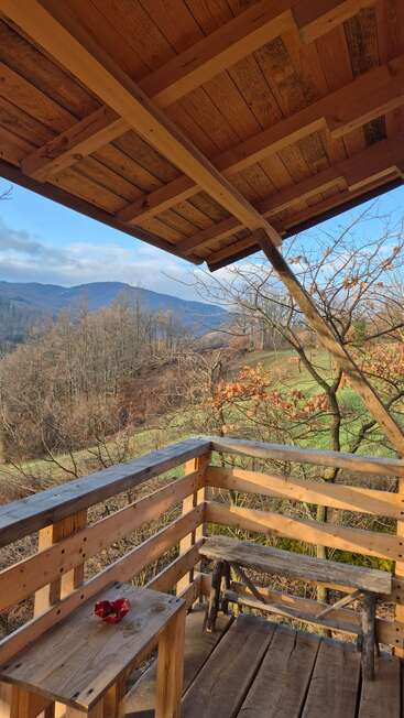 Eine gemütliche Holzveranda bietet einen Blick auf eine malerische Landschaft mit sanften Hügeln, kahlen Winterbäumen und einem strahlend blauen Himmel. Eine rustikale Bank und ein Tisch laden zum Entspannen ein.