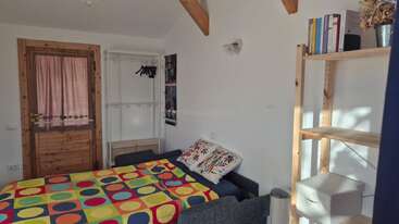 Dieses gemütliche Schlafzimmer hat eine bunte Bettdecke mit Polka-Dots, ein kleines Bücherregal aus Holz mit Büchern, Wandhaken mit Kleiderbügeln, eine Holztür und viel Tageslicht, das hereinströmt.