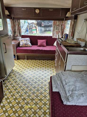 This cozy vintage caravan features maroon seating, patterned curtains, a compact kitchenette, retro fridge, patterned linoleum floor, overhead storage, throw pillows, blankets, and natural light.