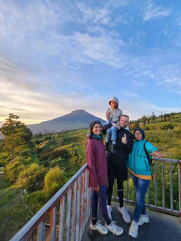 Uma família feliz de quatro pessoas posa em uma plataforma de observação panorâmica, cercada por colinas verdes e uma montanha sob um lindo céu azul com nuvens dispersas.