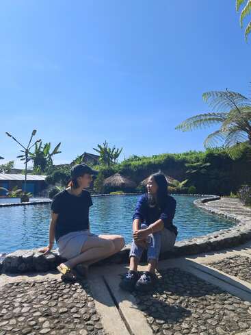 Duas pessoas sentadas à beira de uma bela piscina, aproveitando um dia ensolarado sob um céu azul claro, cercadas por uma vegetação exuberante e plantas tropicais, sorrindo e conversando.
