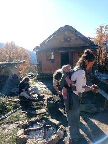 Three adults and a baby work outdoors near a rustic house. The scene features autumn trees, a firepit, and sunlit mountains creating a peaceful, communal atmosphere.