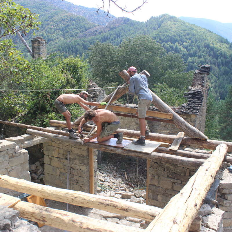 Three shirtless men work on a stone building with wooden beams, surrounded by trees and mountains in the background on a sunny day.