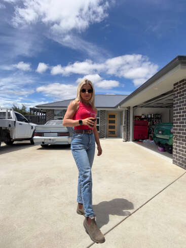 Eine Frau mit Sonnenbrille, rotem, ärmellosem Oberteil, blauen Jeans und Stiefeln steht selbstbewusst vor einem modernen Haus, ihr Handy in der Hand, unter einem teilweise bewölkten Himmel.
