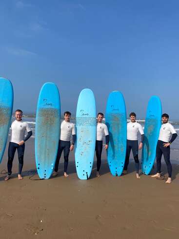 Five men in wetsuits stand on a sandy beach, each holding tall blue surfboards upright. The sky is clear and the ocean is visible behind them.