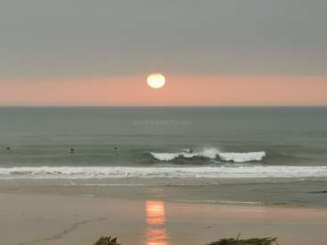 A serene beach at sunset with a glowing sun reflecting on wet sand. Surfers catch gentle waves, creating a peaceful, picturesque scene under a soft, cloudy sky.