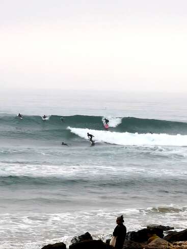 Surfers ride the waves in a misty ocean while a person stands on rocks watching. The sky is overcast, creating a calm, serene atmosphere.