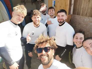 A group of smiling people in white surf shirts pose for a cheerful selfie, standing together in bright sunlight outside a wooden building, ready for surfing.