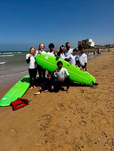 A group of people in matching shirts pose with green surfboards on a sandy beach. The ocean and an old stone building are visible behind them.