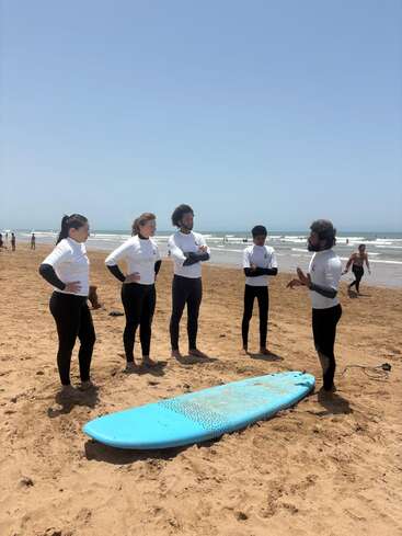 Five people in wetsuits stand on a sandy beach, listening to an instructor beside a blue surfboard, preparing for a surf lesson under a clear sky.