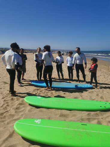 A group of people gathers on a sandy beach, standing by surfboards, wearing wetsuits, preparing for a surfing lesson under a clear, sunny blue sky.