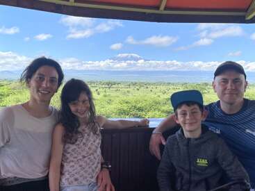 Une famille de quatre personnes souriante pose à l'intérieur d'un véhicule de safari, avec en arrière-plan un paysage verdoyant et le mont Kilimandjaro enneigé visible au loin sous un ciel bleu.