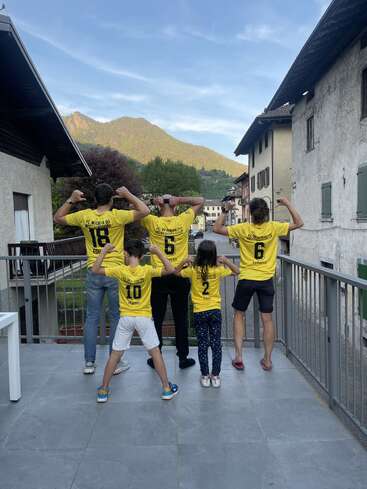 Cinq personnes se tiennent sur une terrasse, dos à la caméra, exhibant leurs muscles dans des maillots jaunes assortis du FC Wacker 90, avec en arrière-plan un paysage de montagnes et de villages.