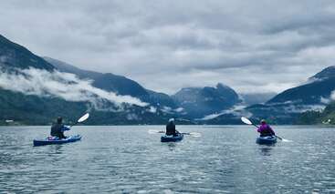 Tres personas navegan en kayak por un tranquilo lago rodeado de montañas neblinosas y boscosas bajo un cielo nublado, creando un ambiente de tranquilidad y aventura al aire libre en la naturaleza.