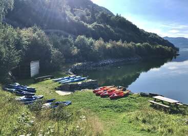 Coloridos kayaks descansan en la orilla cubierta de hierba de un lago sereno, rodeado de exuberantes colinas verdes, árboles y la luz del sol, con mesas de picnic y tranquilos reflejos. Un entorno tranquilo.