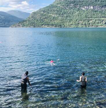 Tres personas disfrutan de un refrescante baño en un lago azul claro, rodeado de exuberantes montañas verdes bajo un cielo parcialmente nublado, creando una tranquila escena veraniega.