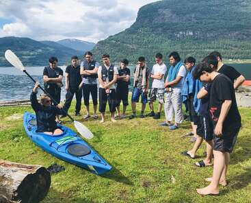Un grupo de adolescentes observa atentamente a un instructor que hace una demostración de técnicas de piragüismo en un kayak azul junto a un pintoresco lago rodeado de verdes colinas y montañas.