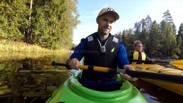 La imagen muestra a un hombre en un kayak verde sosteniendo un remo, con otra persona en un kayak amarillo detrás de él, en un río o lago rodeado de árboles.