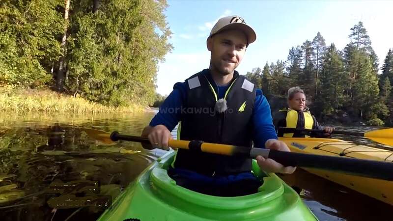 La imagen muestra a un hombre en un kayak verde sosteniendo un remo, con otra persona en un kayak amarillo detrás de él, en un río o lago rodeado de árboles.