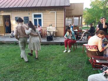 A group enjoys an outdoor gathering with people dancing, playing musical instruments, and relaxing near a cozy wooden house on a sunny day.