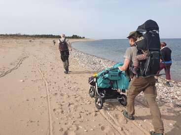 A group of people hiking along a sandy beach, pushing a stroller and wearing backpacks. The shoreline is pebbly, with calm sea and distant greenery.