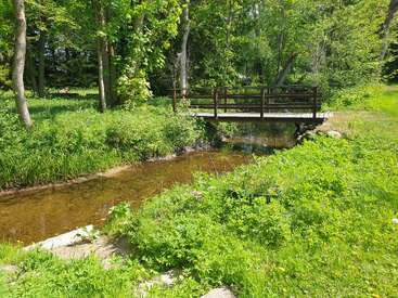 A small wooden bridge crosses a clear, shallow stream surrounded by lush greenery and tall trees, creating a peaceful, sunny, and inviting natural scene.