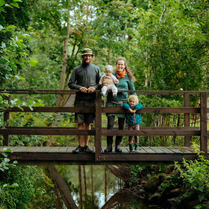 A smiling family of four stands on a wooden bridge in a lush green forest, enjoying nature together. Two young children complete the happy scene.