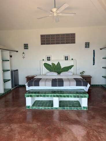 Um quarto aconchegante com uma cama grande, piso de azulejos verdes, decoração minimalista, ventilador de teto, cabeceira com folhas, mesinhas laterais, prateleiras abertas e piso em tom de vermelho terroso.