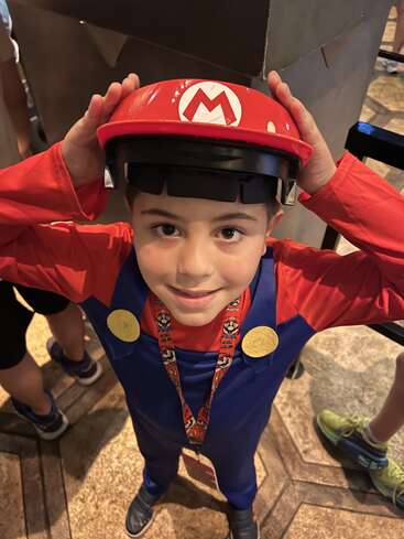 Uma criança sorridente fantasiada de Mario, do Super Mario, usa um boné vermelho e um macacão azul, olhando para cima enquanto segura a aba do boné, em pé dentro de casa com outras pessoas ao redor.