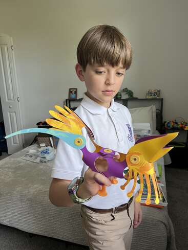 Um menino vestindo uma camisa polo branca e calças cáqui está segurando um modelo colorido e abstrato de um pássaro em um quarto arrumado e bem iluminado.