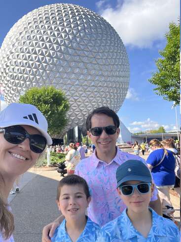 Uma família sorridente de quatro pessoas posa em frente à icônica esfera geodésica do Epcot, vestindo roupas combinando em azul e rosa, aproveitando um dia ensolarado na Disney.