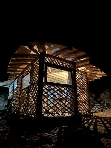 Une cabane confortable en bois avec des murs en treillis est éclairée de l'intérieur et se dresse seule dans la nuit noire, projetant des ombres complexes sur le sol à l'extérieur.