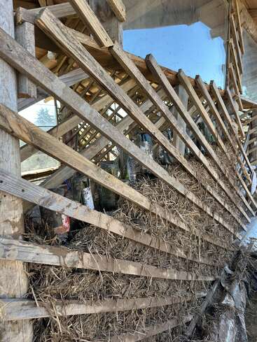 L'image montre une structure en treillis en bois remplie de paille et de bouteilles en verre, formant un mur rustique et durable. La lumière du soleil se reflète sur le verre, créant une esthétique terreuse.