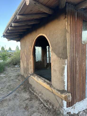 This image shows a rustic, unfinished adobe building with a circular window, wooden beams, corrugated metal, and a dirt path surrounded by vegetation under a blue sky.