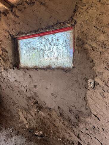 This image shows a partially finished window in a mud or adobe wall, with rough earthen texture surrounding the glass and an unfinished electrical outlet.