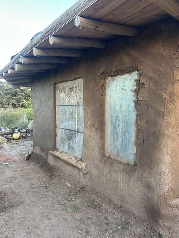 This image shows a rustic adobe house with a wooden roof, two covered windows, and an earthy wall. Outdoor tools and greenery are visible nearby.