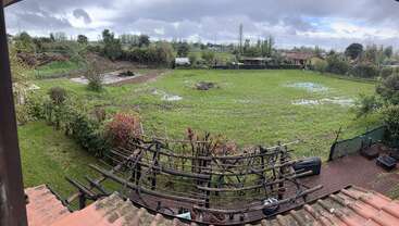 This image shows a large, grassy backyard with patches of water, a wooden pergola, garden plants, a patio, and a cloudy sky above. Peaceful countryside scene.