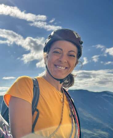 A smiling woman wearing a helmet and yellow shirt stands outdoors, with a backpack and rope, against a scenic mountain backdrop under a bright blue sky.