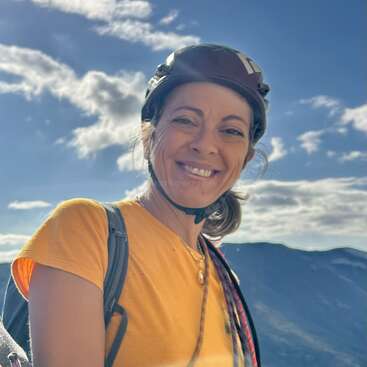 A smiling woman wearing a helmet and yellow shirt stands outdoors, with a backpack and rope, against a scenic mountain backdrop under a bright blue sky.