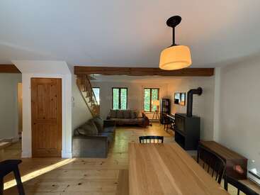 Dieser gemütliche Wohnbereich verfügt über Holzböden, sichtbare Balken, einen Holzofen, bequeme Sofas, einen Esstisch, ein Klavier, große Fenster und eine warme, einladende Atmosphäre.