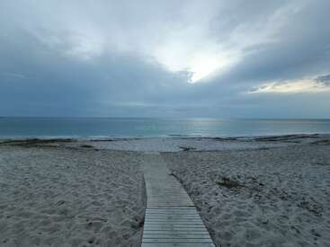Das Bild zeigt eine ruhige Strandszene mit einer hölzernen Strandpromenade, die zum Meer führt, vor einem wolkenverhangenen Himmel mit der Sonne, die durch die Wolken blickt.