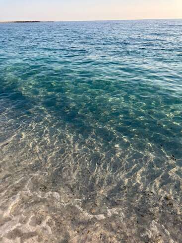 Klares türkisfarbenes Wasser umspült sanft einen Sandstrand unter einem ruhigen, hellblauen Himmel. Das Sonnenlicht erzeugt schimmernde Muster auf dem Meeresgrund und ruft Frieden und Ruhe hervor.