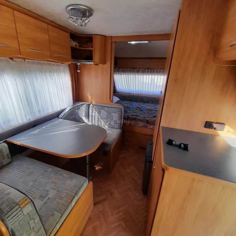Das Bild zeigt das Innere eines kompakten Wohnmobils mit Essbereich, Sitzgelegenheiten und Schlafplätzen, mit Holzverkleidung und einer kleinen Küchenzeile.