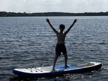 Un jeune garçon se tient triomphalement sur un paddleboard au milieu d'un lac, les bras levés en signe de joie, l'eau scintillante, avec des arbres en arrière-plan.