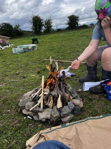 Un feu de camp construit à partir de bâtons et entouré de pierres brûle en plein air. Une personne portant des bottes et un masque coloré est assise à proximité, sous un ciel nuageux.