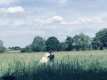 Une personne et un grand chien blanc s'étreignent chaleureusement dans une prairie verdoyante, sous un ciel bleu parsemé de nuages cotonneux et d'arbres aux couleurs vives.