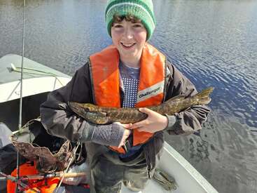 Un garçon souriant, coiffé d'un bonnet vert et vêtu d'un gilet de sauvetage orange, tient un poisson fraîchement pêché, debout dans une petite barque sur un lac calme.
